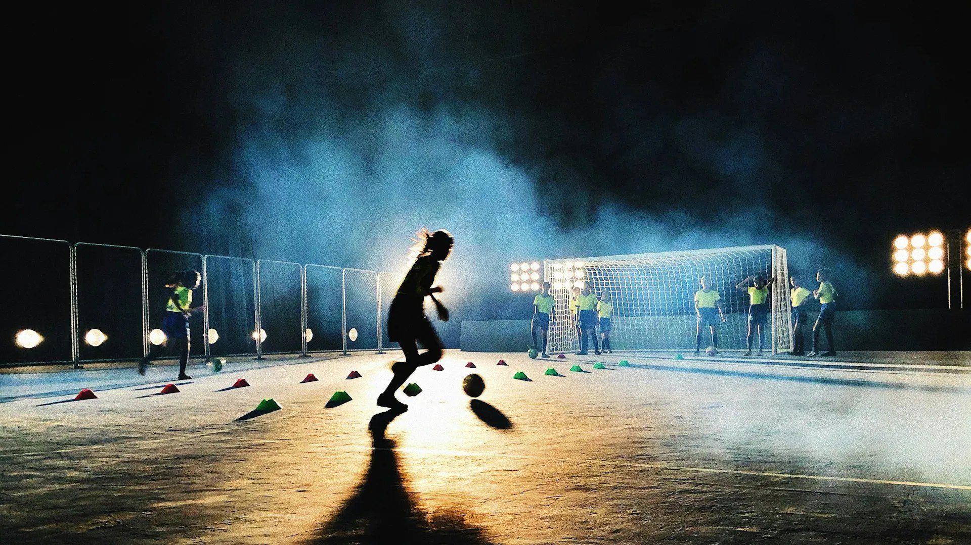 A young player dribbling through cones under floodlights